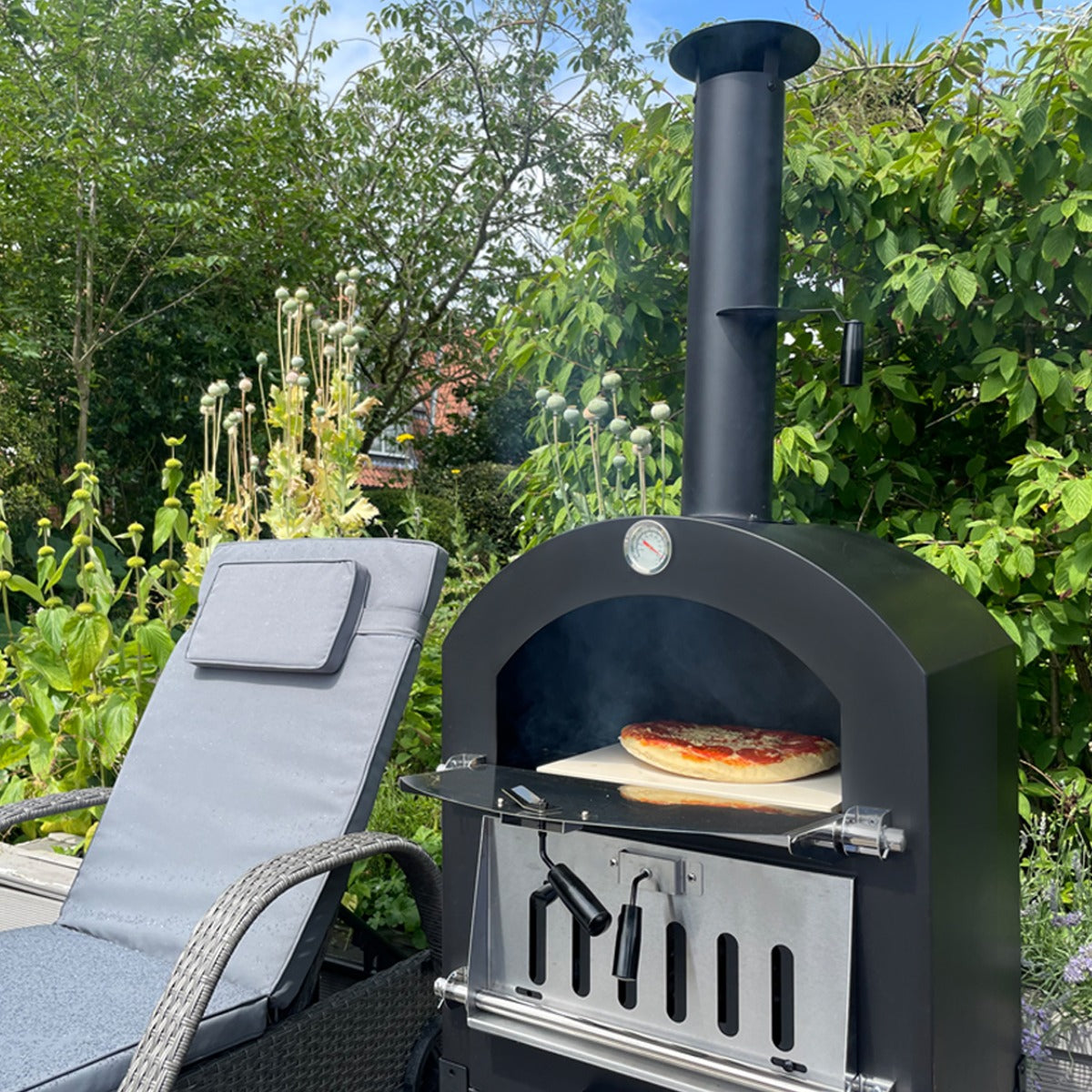 Schwarzer KuKoo Outdoor Pizzaofen mit Holzfeuer, auf dem eine frische Pizza backt, umgeben von grünen Pflanzen und einem gemütlichen Stuhl