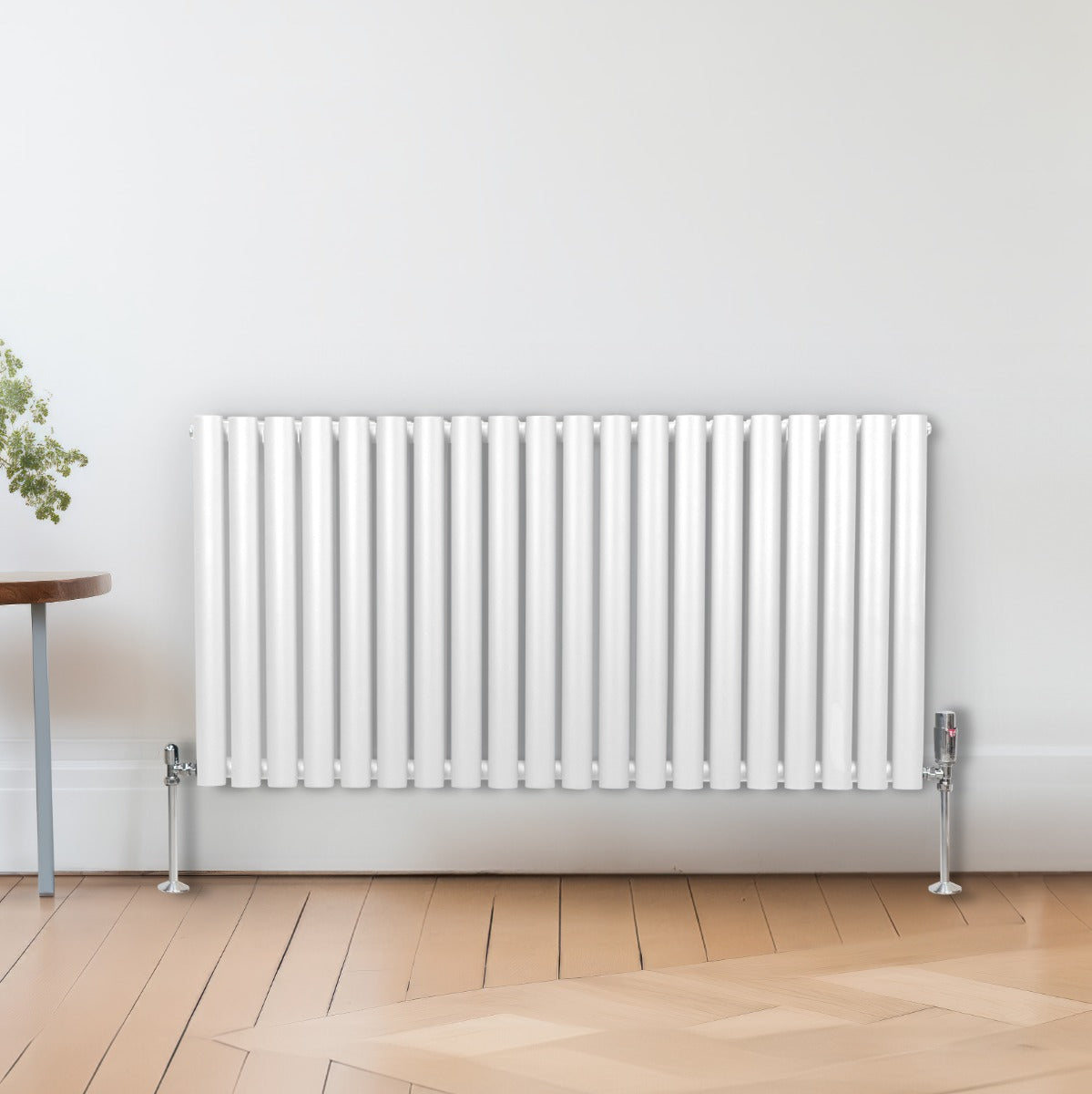 Ovaler Röhrenheizkörper in Weiß mit Chrom-Thermostatventilen, 600mm x 1200mm, auf einem hellen Holzboden, umgeben von einer Pflanze und einem Tisch