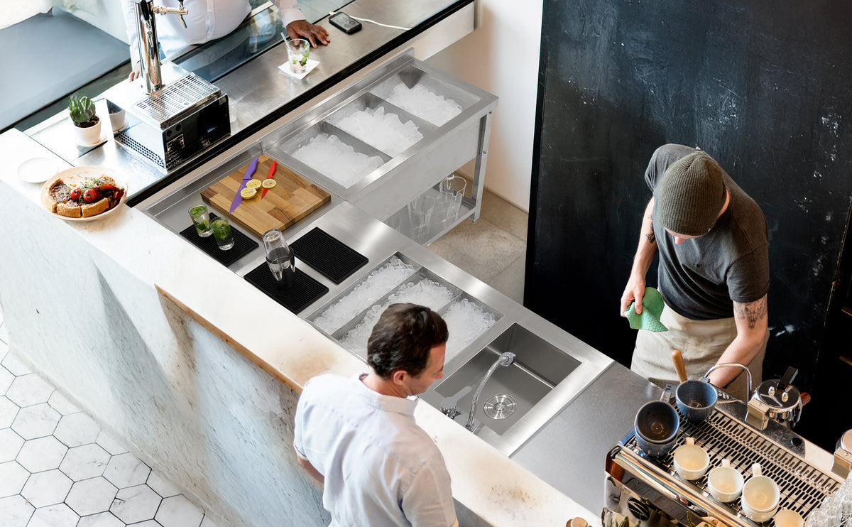 Moderne Café-Theke mit Edelstahloberfläche, Eisbehältern, frischen Zutaten und Barista, der Getränke zubereitet, im Hintergrund eine gemütliche Atmosphäre
