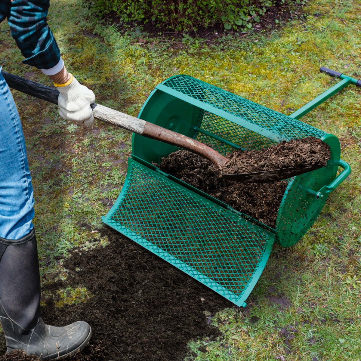 Grüner Garten Kompoststreuer aus Metall, mit grobem Netz, wird mit einer Schaufel gefüllt, während frischer Kompost auf den Boden verteilt wird