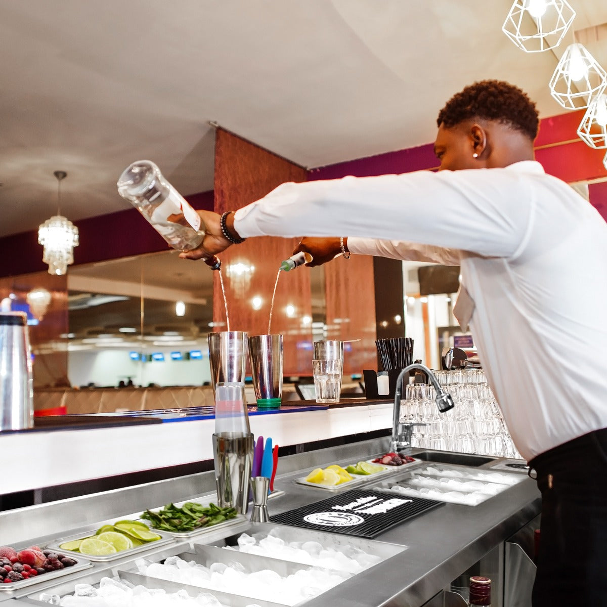 Ein Barkeeper mixt Cocktails an einer modernen Duo-Barstation mit Edelstahloberfläche, frischen Zutaten und Eiswürfeln in Gläsern