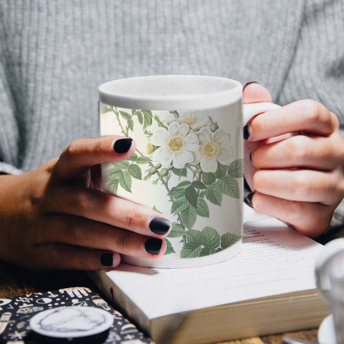 Weißer Becher mit floralen Motiven, gehalten von einer Person mit schwarzen Nägeln, auf einem Holztisch neben einem Buch und einer Tasse