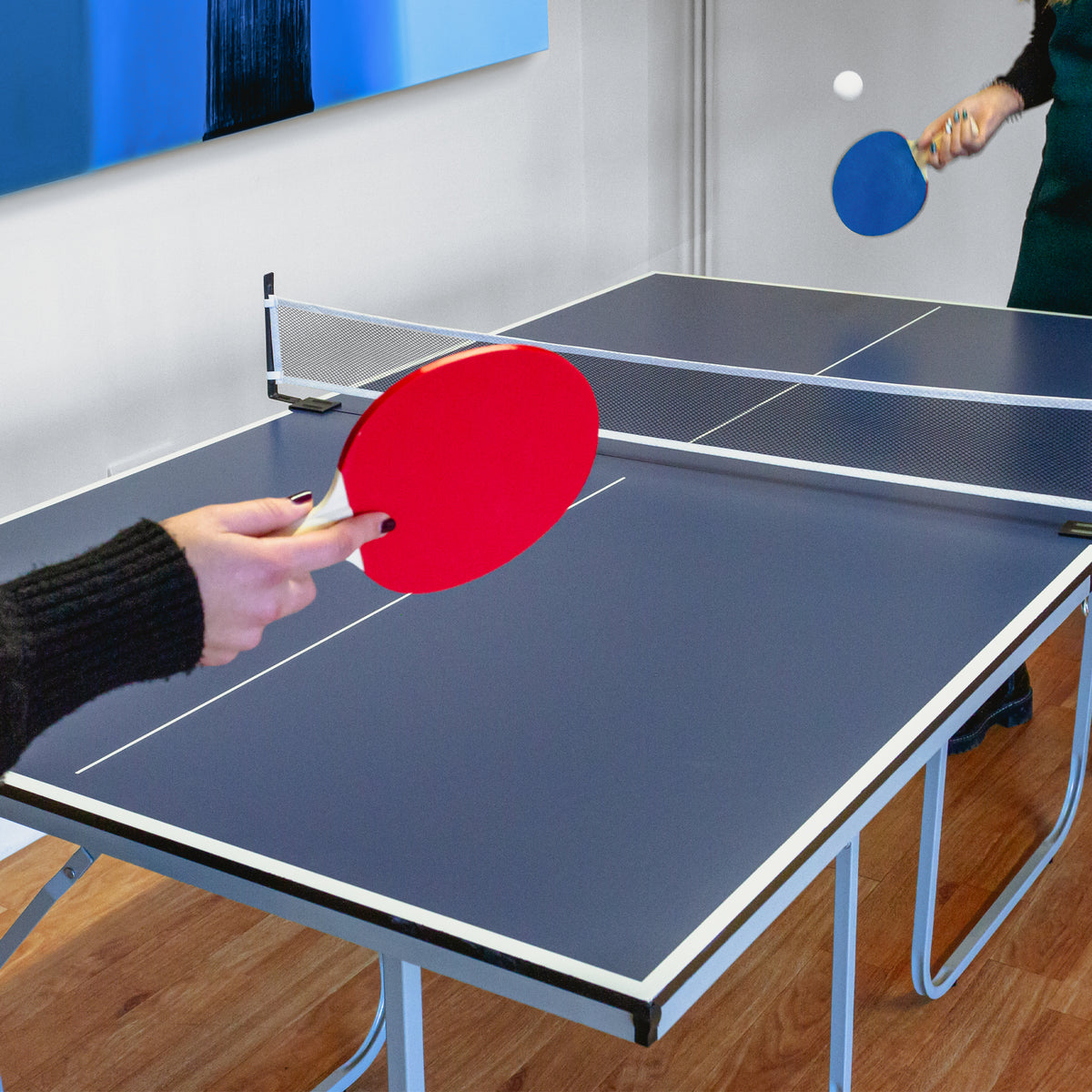 Tischtennisplatte in Blau mit Netz, zwei Spieler halten rote und blaue Schläger, Holzfußboden im Hintergrund, zwei zusätzliche Tischtennisbälle sichtbar