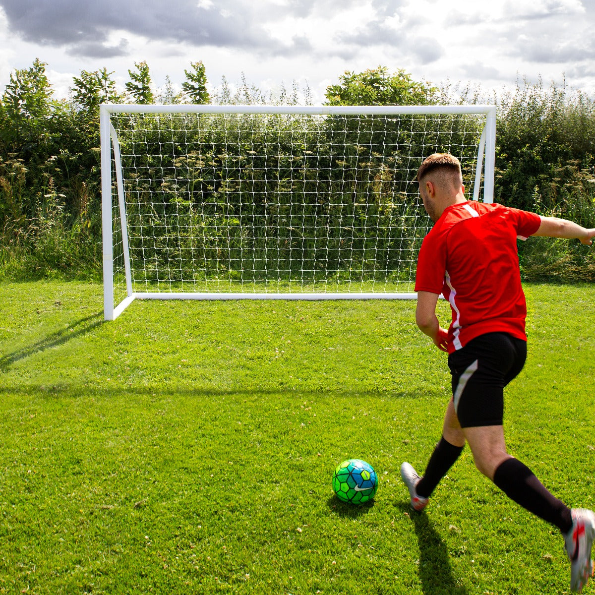 Fußballtor in Weiß mit Netz, 3,6m x 1,8m, auf grünem Rasen, Spieler in rotem Trikot schießt auf einen bunten Fußball, Wolken am Himmel