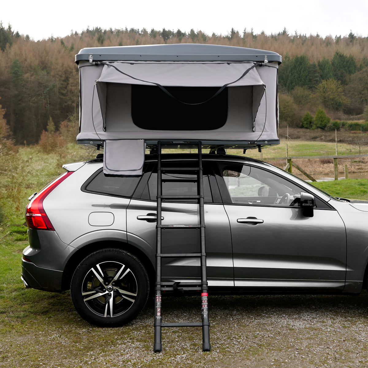 Dachzelt auf einem grauen SUV, mit einer stabilen Leiter, umgeben von grüner Wiese und Bäumen im Hintergrund, ideal für Campingabenteuer