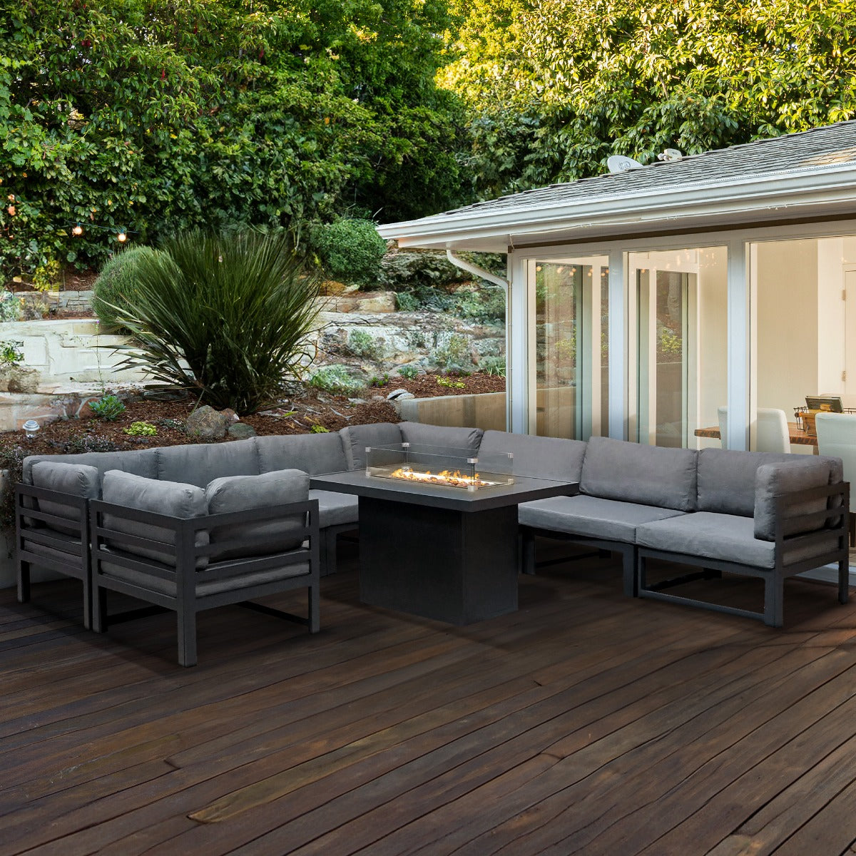 Modulares Gartenlounge-Set mit grauen Polstern und integrierter Feuerstelle, umgeben von grünem Garten und Holzterrasse