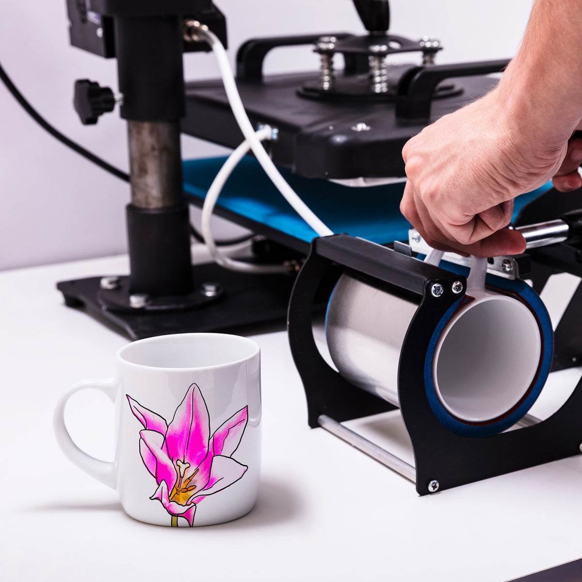 Weißer Sublimationsbecher mit pinker Orchideenblüte, daneben eine Heizbandrolle und eine Druckmaschine auf einem weißen Tisch