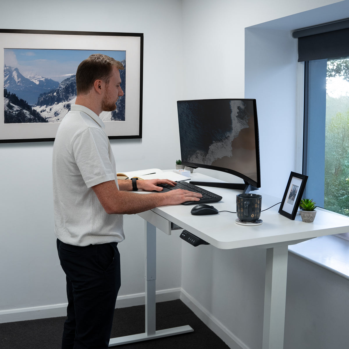Ein Mann steht an einem höhenverstellbaren Schreibtisch, arbeitet am Computer, umgeben von modernen Büromöbeln und einer Berglandschaft im Hintergrund