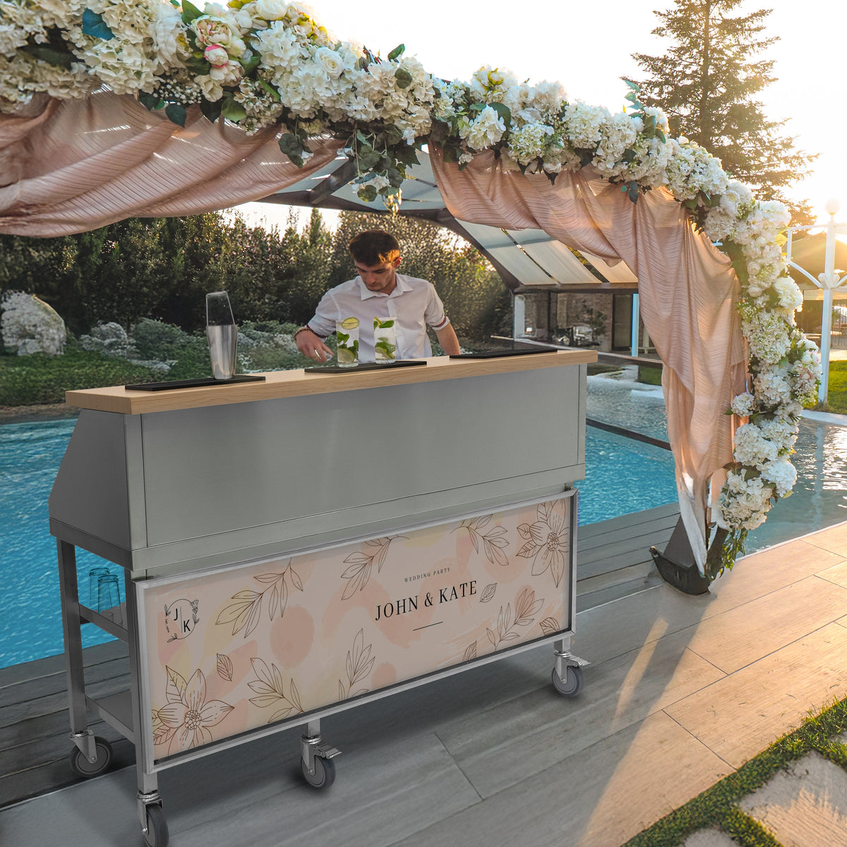 Mobile Cocktailbar Station in sanftem Grau mit Holzarbeitsfläche, dekoriert mit floralen Akzenten, vor einem Pool und einer romantischen Kulisse