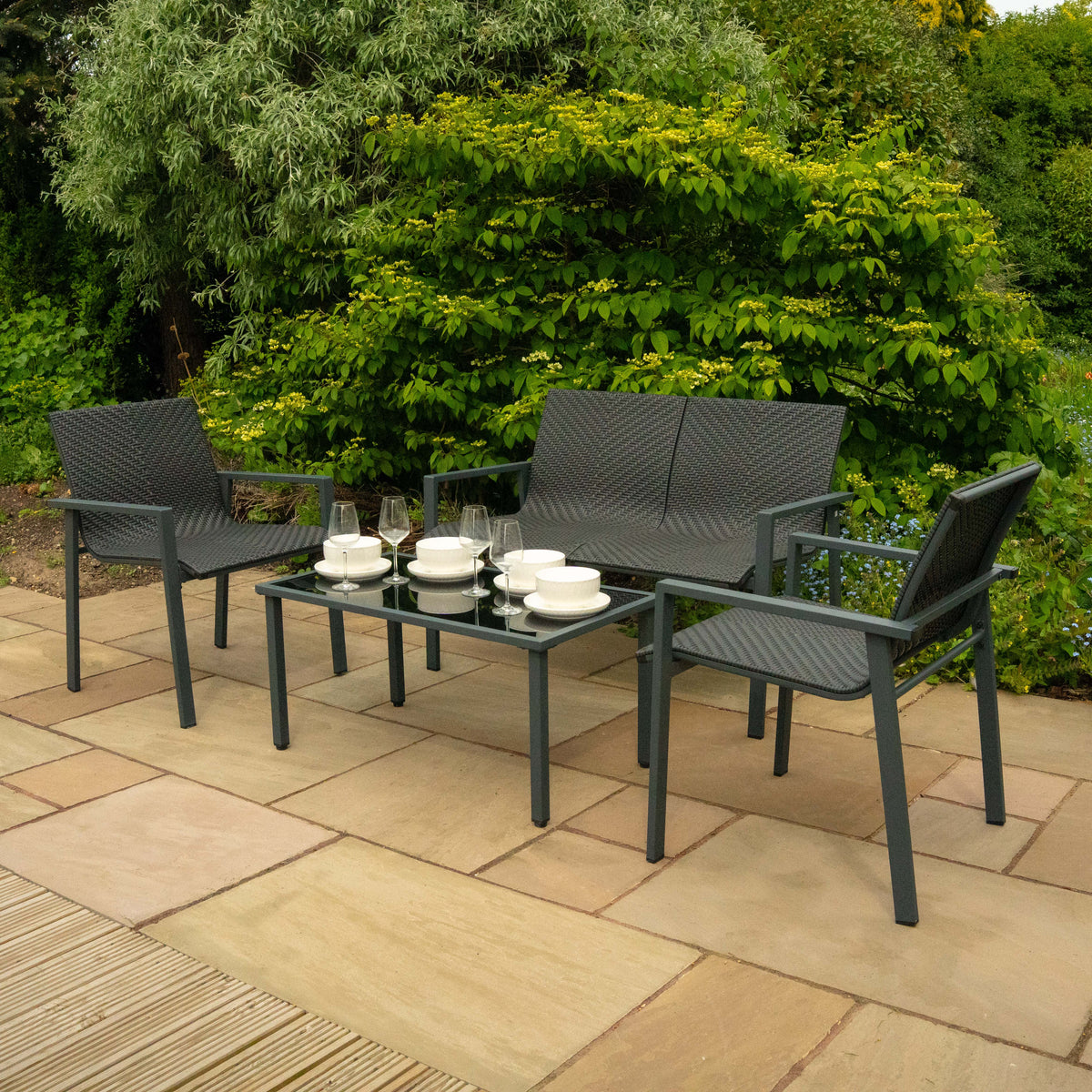 Schwarzes Rattan Terrassenmöbel Set mit zwei Stühlen und einem Tisch, dekoriert mit Geschirr und Gläsern, umgeben von grünen Pflanzen