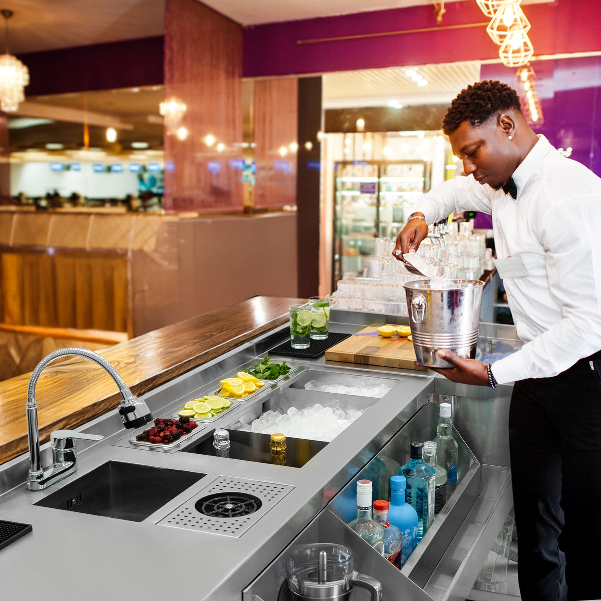 Elegante modulare Deluxe-Cocktailbar-Station mit Eisbrunnen, ausgestattet mit frischen Zutaten, Gläsern und einem Barkeeper in formeller Kleidung