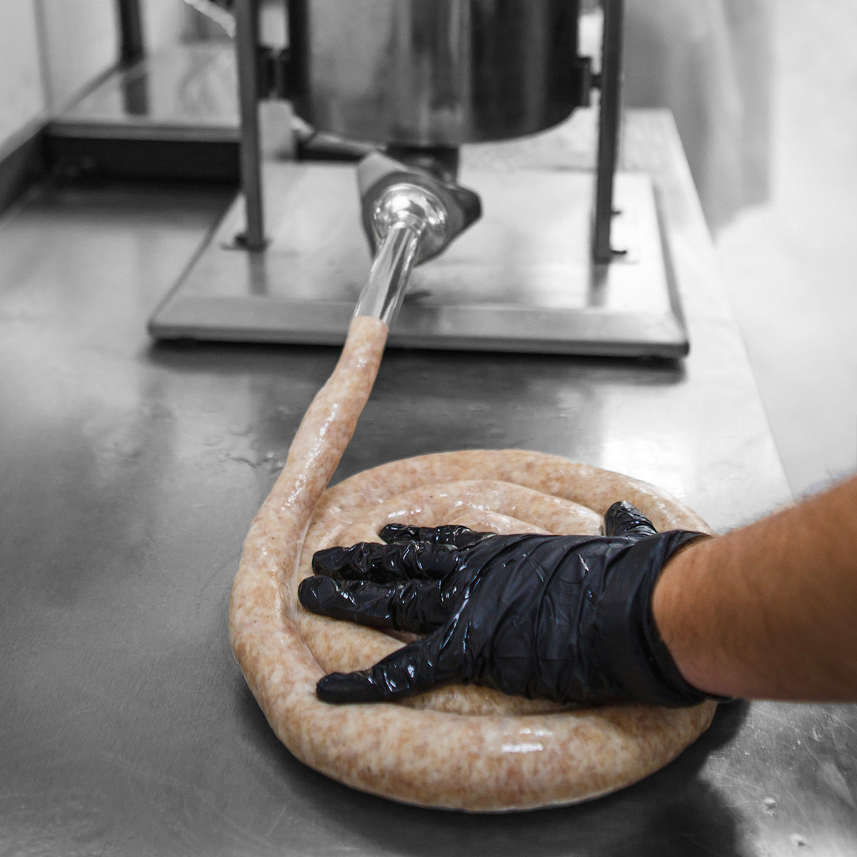 Kukoo Wurstfüller in Aktion, mit einem Handgriff, der frische Wurstmasse auf einem Edelstahl-Tisch formt, umgeben von glänzendem Metall