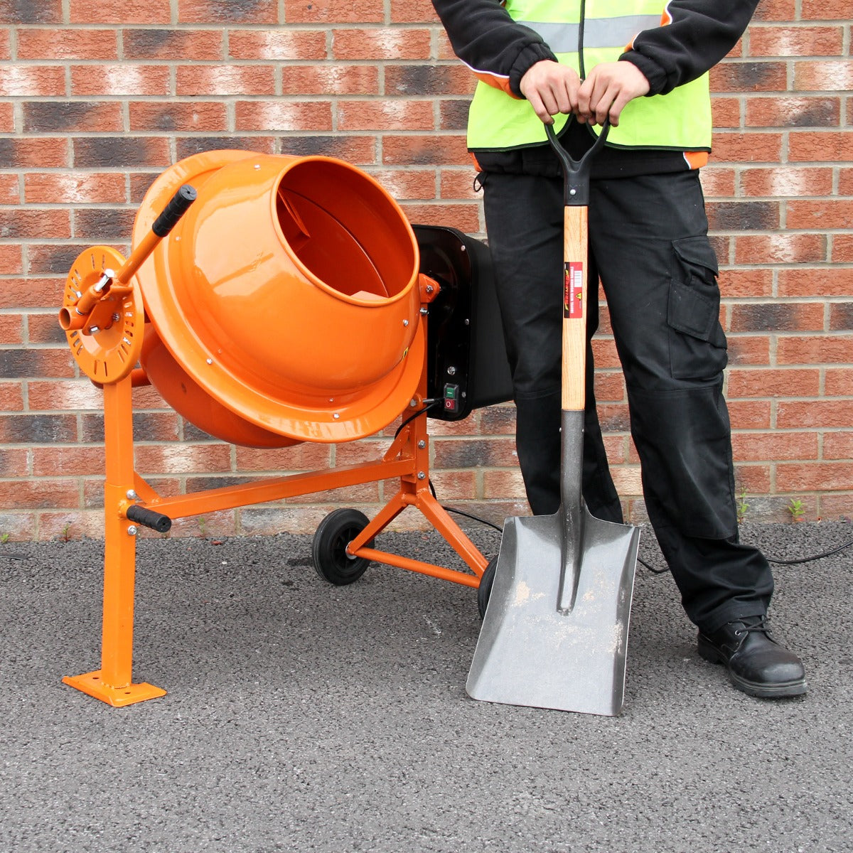 Zementmischer in leuchtendem Orange mit Handbetonrüttler, 2 Meter Schaft, auf grauem Untergrund, neben einer Person in Arbeitskleidung mit Schaufel