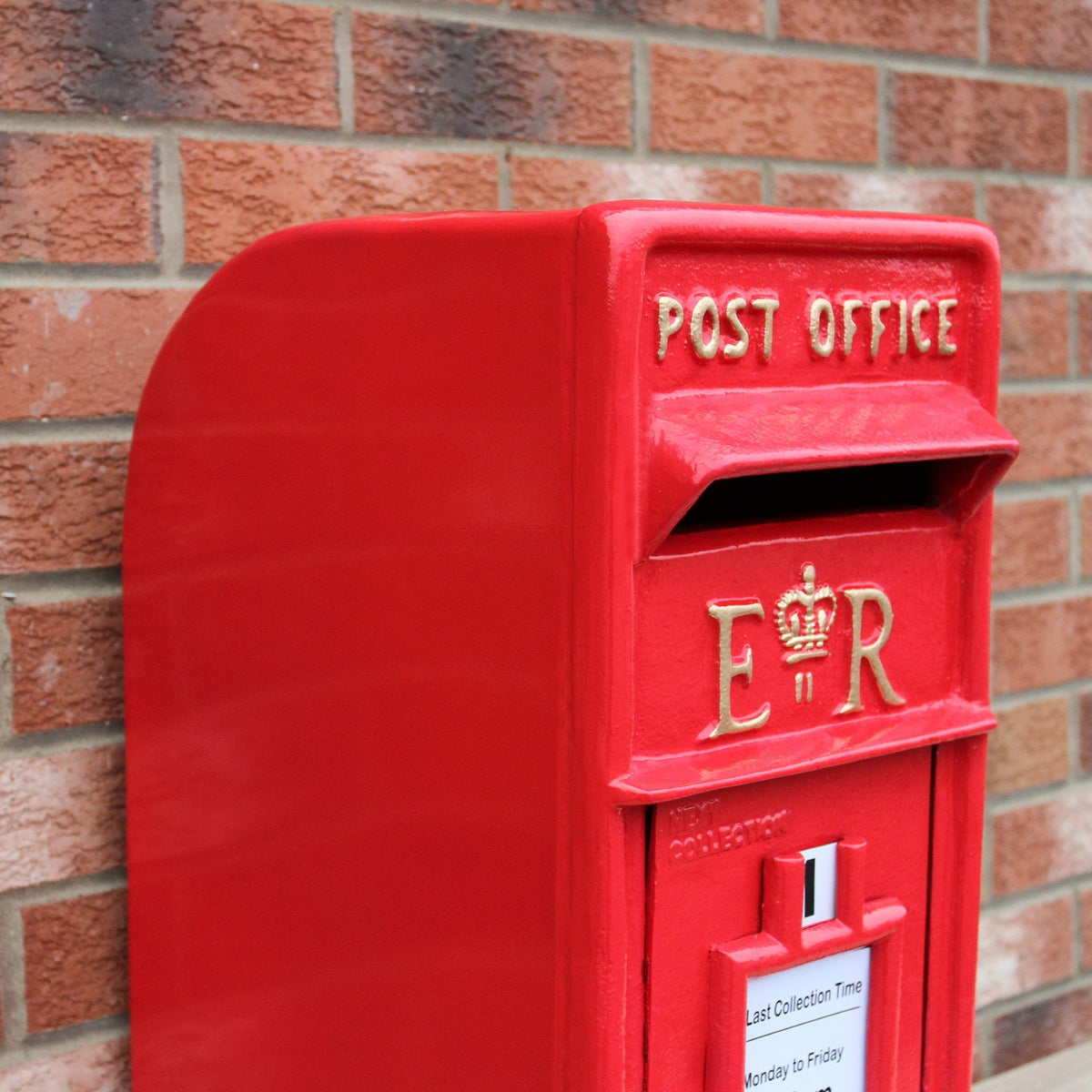 Roter Briefkasten mit Bodenständer, glänzende Oberfläche, goldene Schriftzüge "POST OFFICE" und "ER", vor einer roten Ziegelmauer platziert