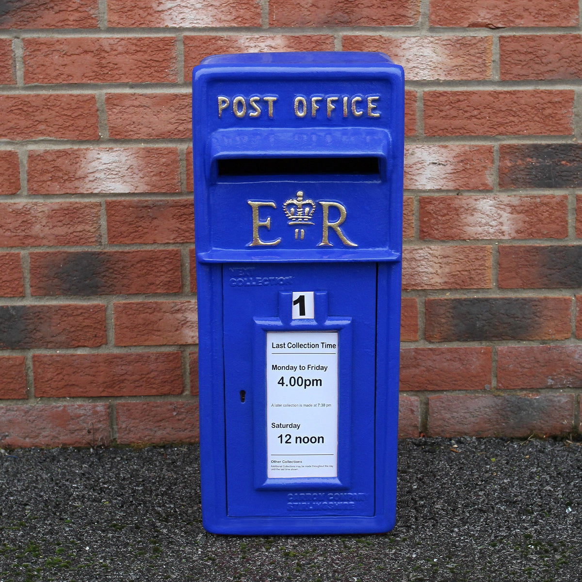 Blauer Briefkasten im schottischen Stil mit goldenen Buchstaben, auf rotem Ziegelhintergrund, zeigt Abholzeiten und königliches Emblem