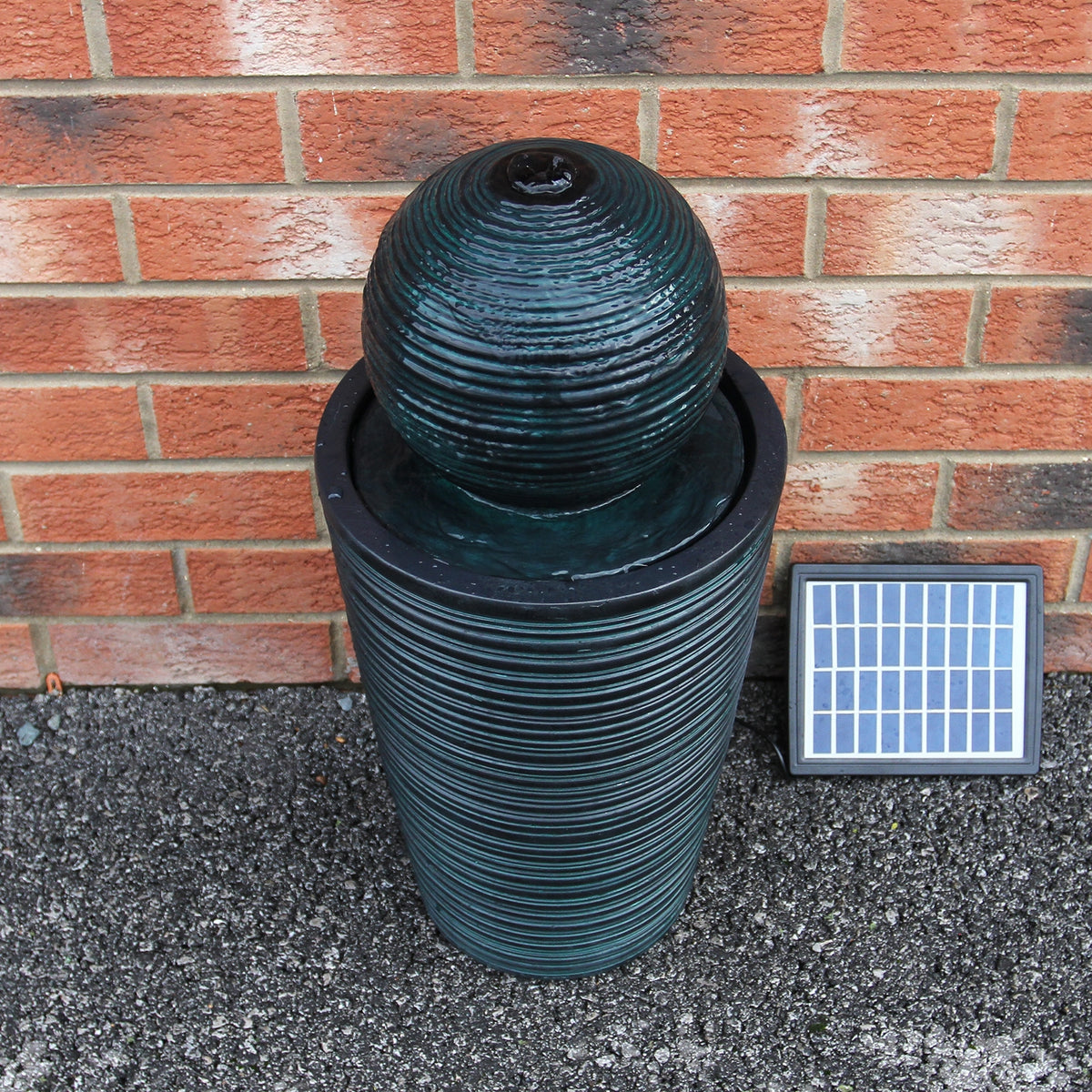 Schwarzer Kugelbrunnen mit strukturiertem, gewelltem Design, umgeben von Ziegelwand und Solar-Panel, ideal für Garten oder Terrasse