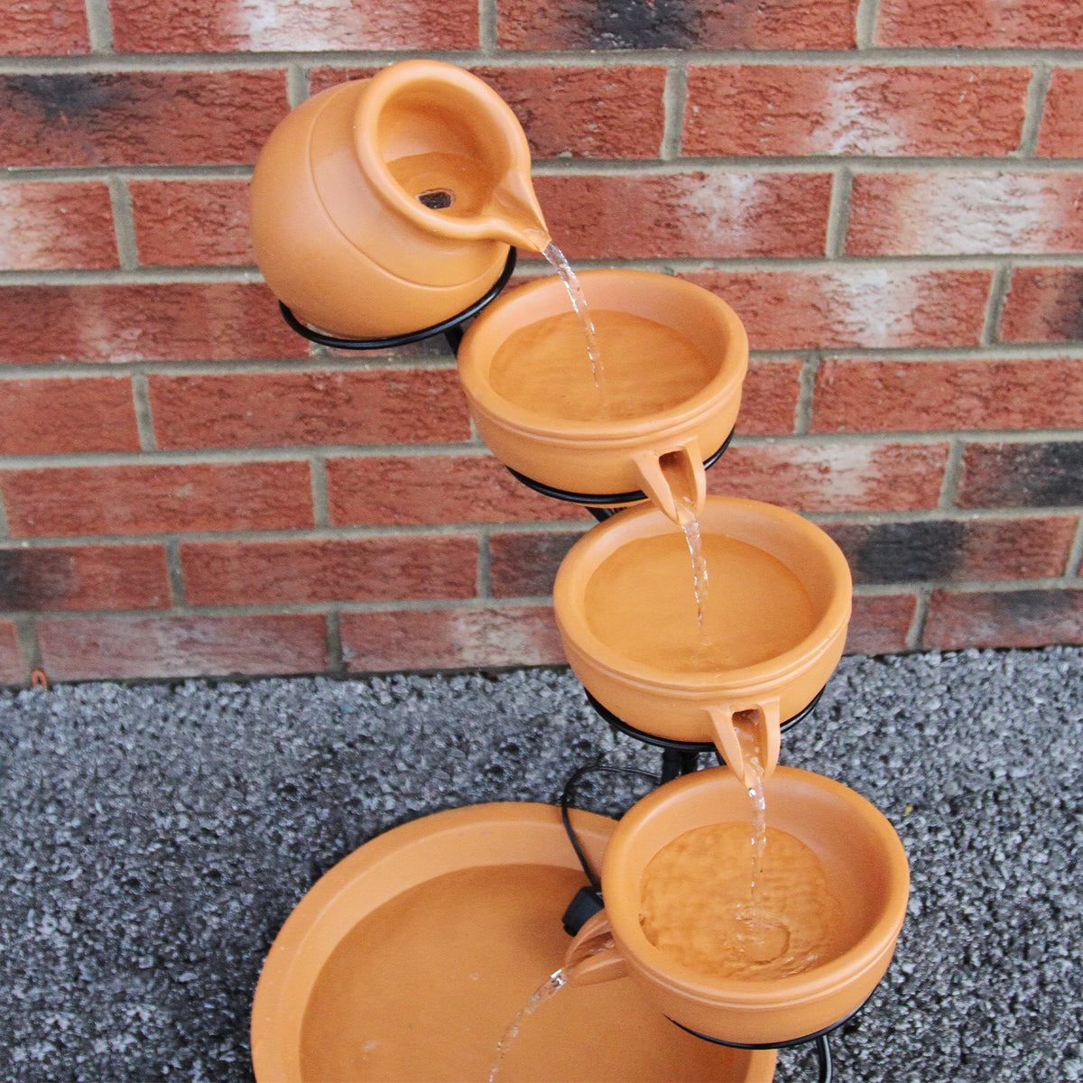Terracotta Solar Kaskadenbrunnen mit vier Schalen, die Wasser in eine große Schale leiten, umgeben von grauem Kies und einer roten Ziegelmauer