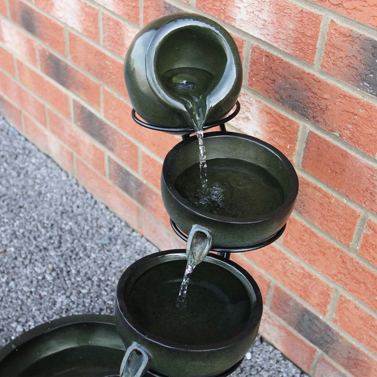 Grüner Solar Kaskadenbrunnen mit Akku, bestehend aus drei Töpfen, die Wasser sanft in eine darunterliegende Schale fließen lassen, vor einer Backsteinwand