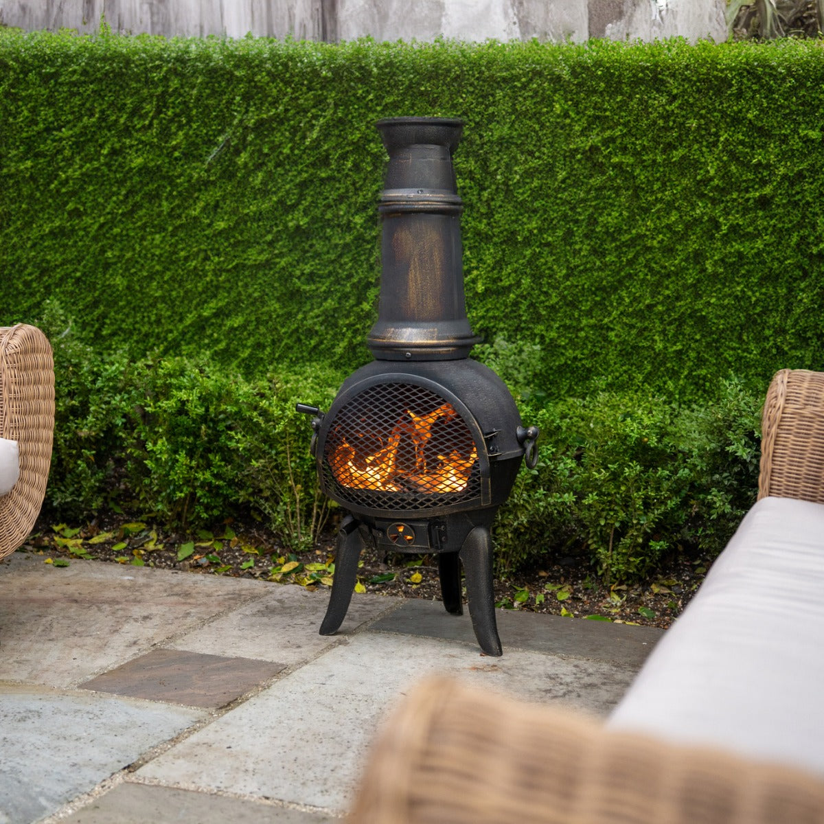 Gusseisen Terrassenofen mit rustikalem Design, schwarzer Oberfläche, rauchigem Kamin und loderndem Feuer, umgeben von grünem Garten und bequemen Sitzmöbeln