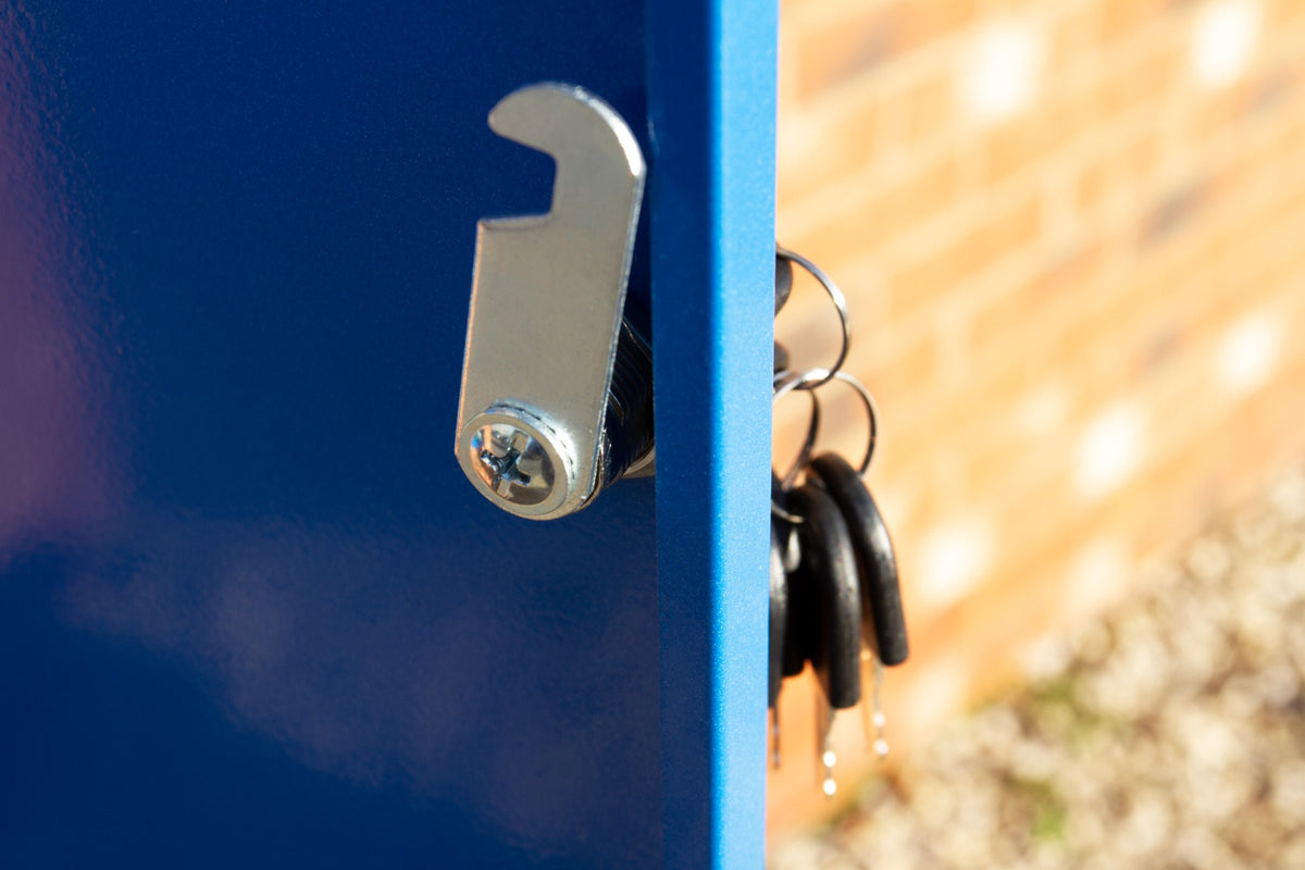 Blauer Paketkasten mit glänzender Metallverriegelung und einem Schlüsselbund, der an der Tür hängt, vor einer gemusterten Backsteinmauer