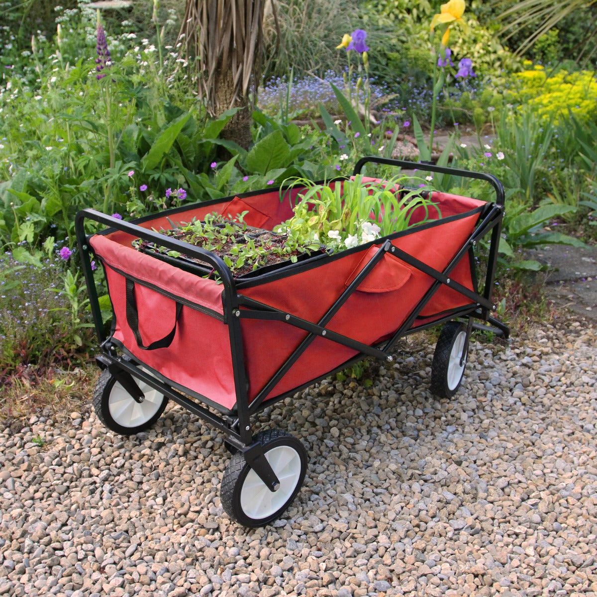 Faltbarer Gartenwagen in leuchtendem Rot mit stabilen schwarzen Rädern, gefüllt mit bunten Pflanzen, steht auf einem Kiesweg umgeben von Blumenbeeten