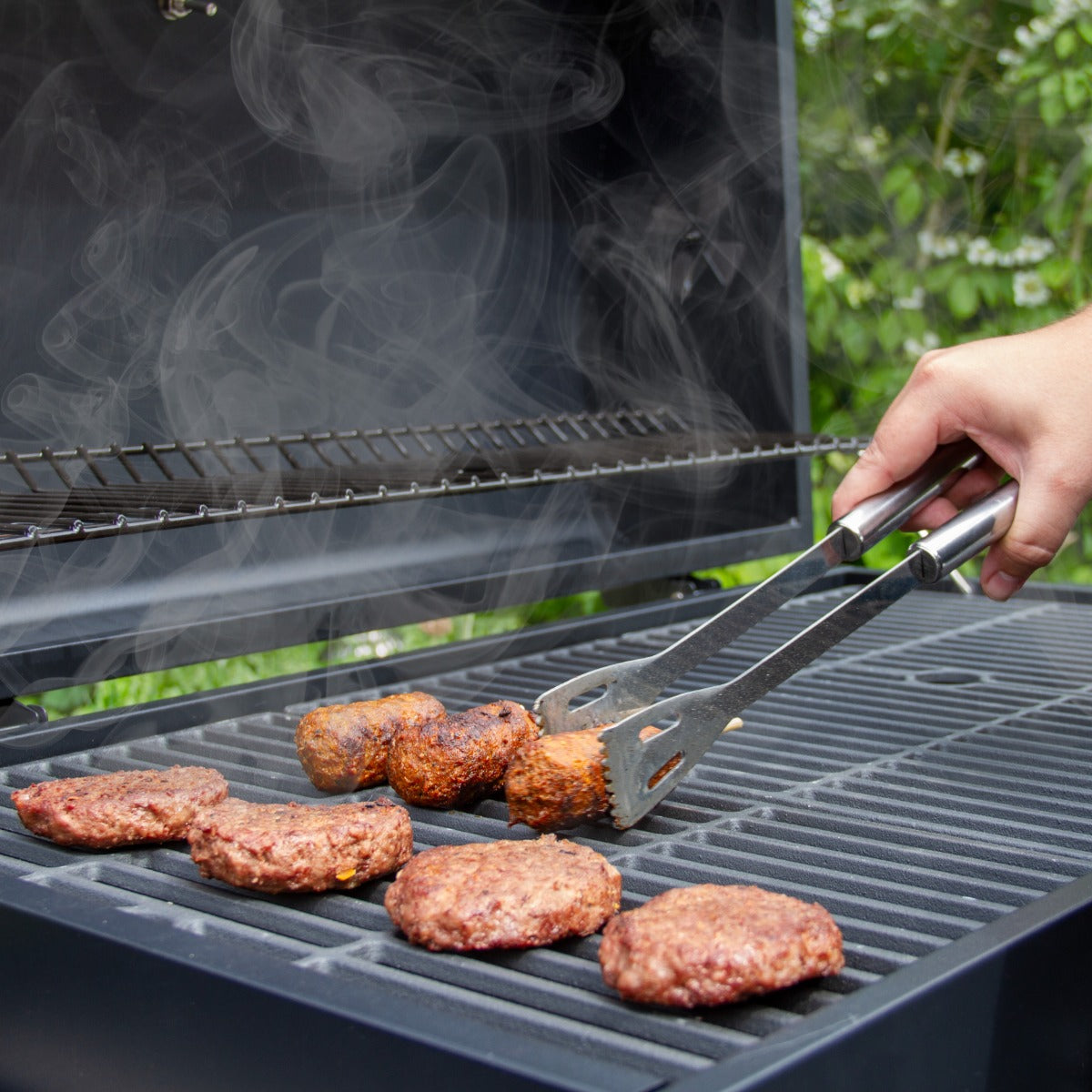 Saftige Burger-Patties brutzeln auf dem Grillrost eines XXL Holzkohlegrills, während Rauch aufsteigt und eine Hand mit Zange das Grillgut wendet