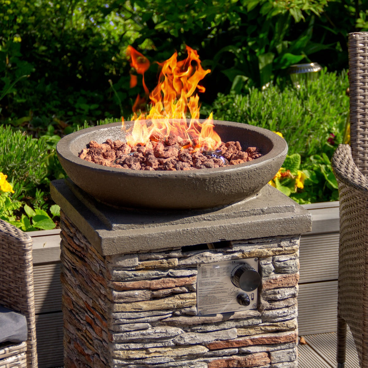 Runde Gas Feuerstelle aus grauem Stein mit flammenden Feuer und Kieselsteinen, umgeben von grüner Vegetation und gemütlichen Gartenmöbeln