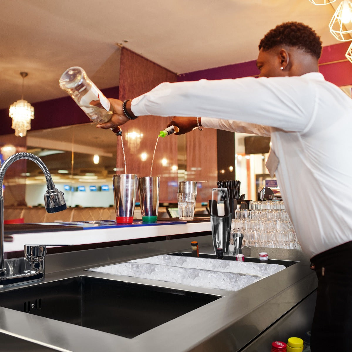 Elegante Cocktail-Bar Station mit einem Barkeeper, der Getränke in Edelstahl-Shaker gießt, umgeben von Gläsern und Eiswürfeln