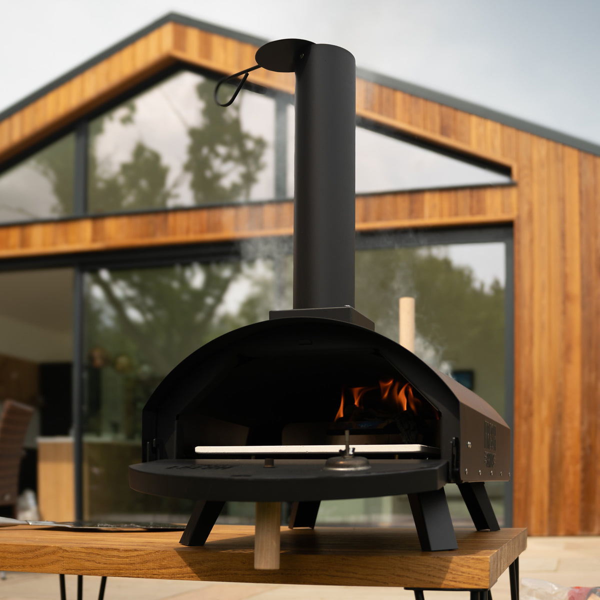 Klassischer Outdoor-Pizzaofen in Schwarz mit einem eleganten, runden Design, Holzfeuerstelle und einem hohen Schornstein, platziert auf einem Holztisch