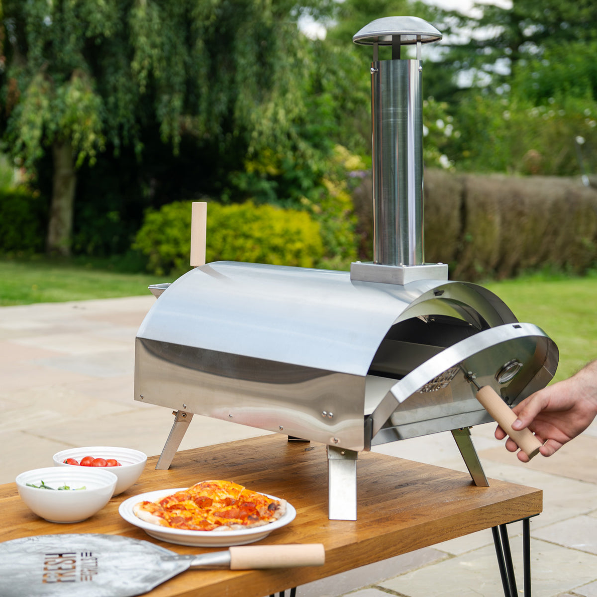 Glänzender, silberner Premium Outdoor-Pizzaofen mit Edelstahl-Oberfläche, auf einem Holztisch, umgeben von frischen Zutaten und einer Pizza