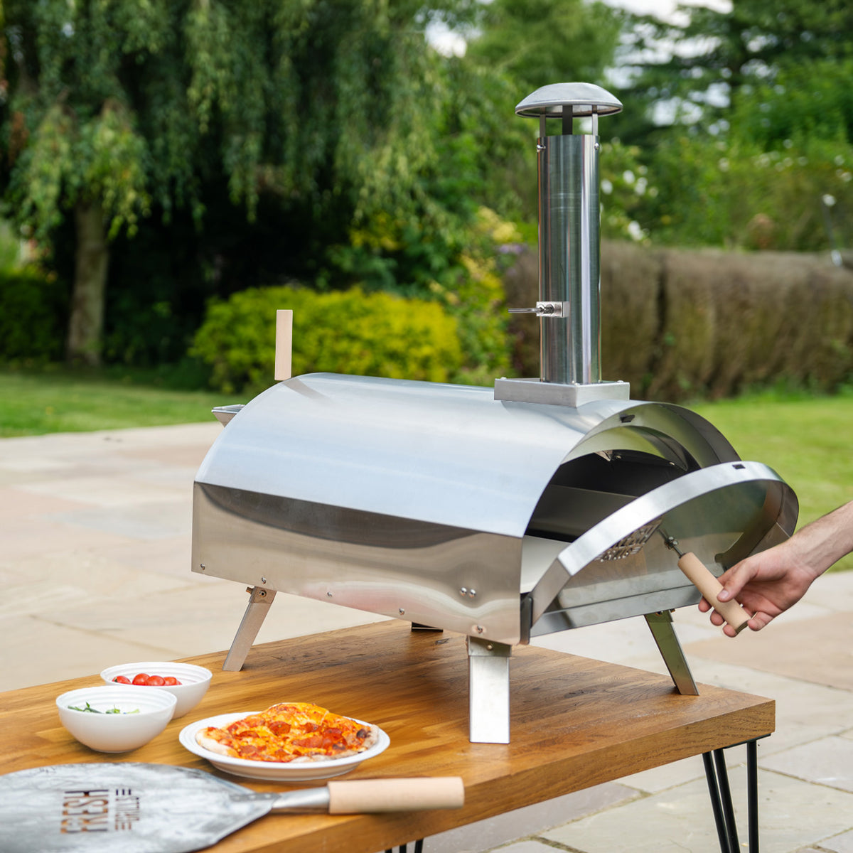 Hochwertiger, extra großer Outdoor-Pizzaofen aus Edelstahl mit glänzender Oberfläche, auf einem Holztisch, umgeben von frischem Gemüse und Kräutern