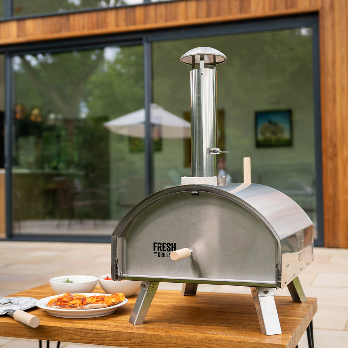 Eleganter, extra großer Outdoor-Pizzaofen aus Edelstahl mit einem runden Design, einem Kamin und Holzgriff, auf einem Holztisch neben frischen Zutaten