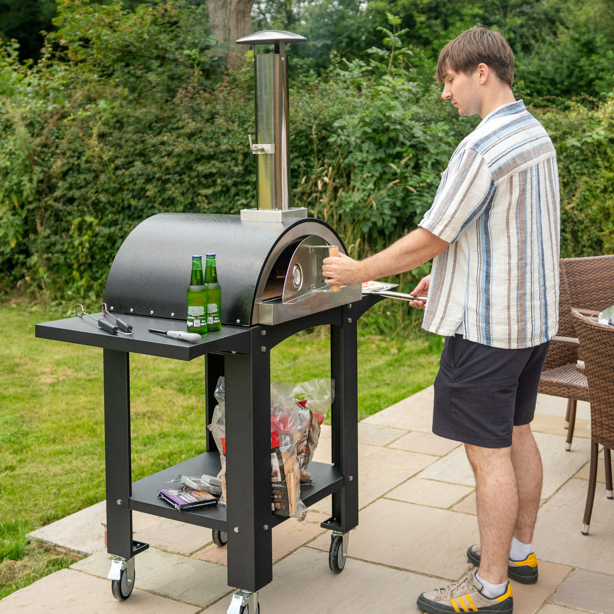 Freistehender Outdoor-Pizzaofen mit schwarzem Gehäuse, Vorbereitungsstation und einem Mann, der Pizza auf einem Holzofen backt, umgeben von Gartenmöbeln