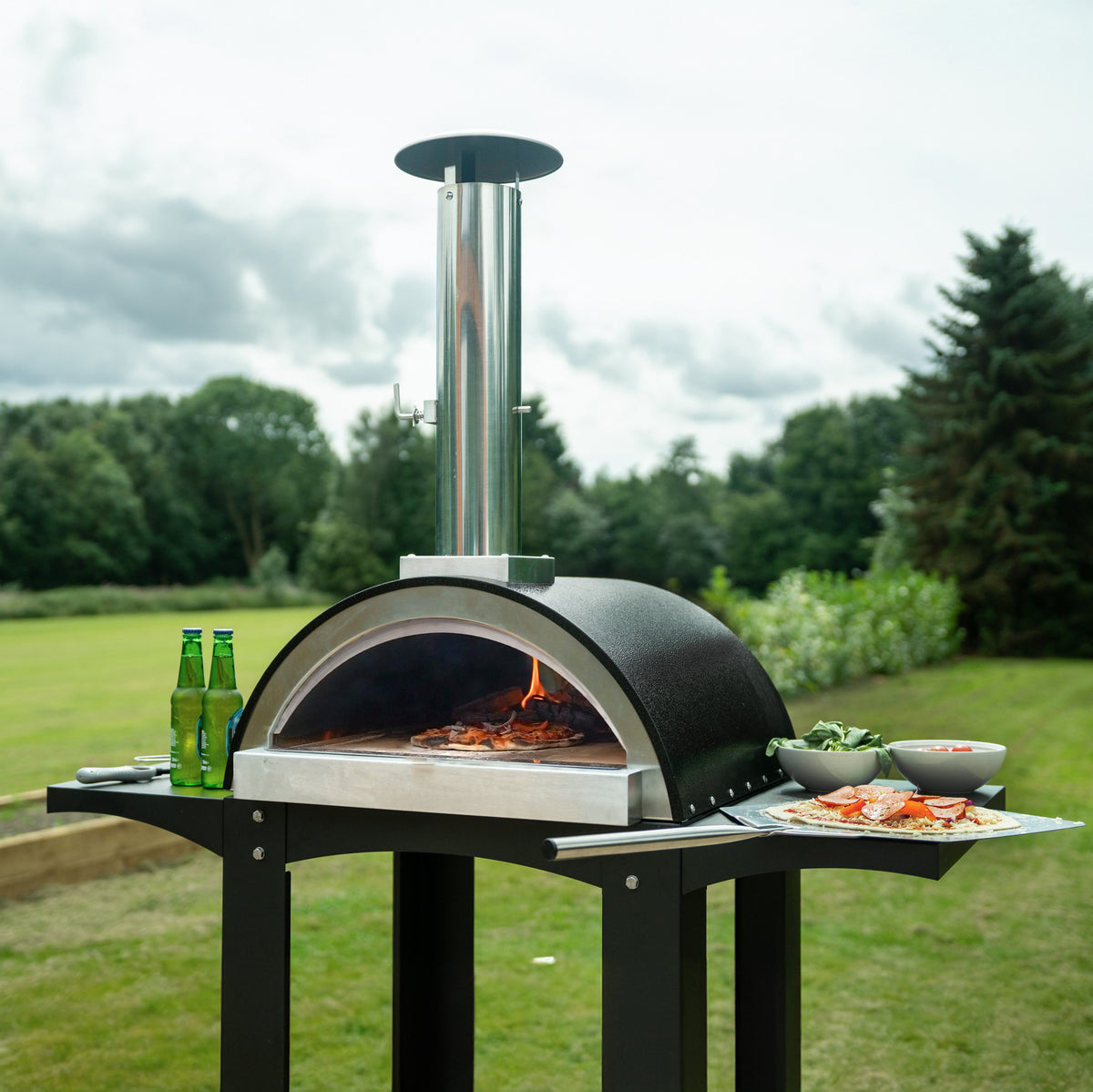 Freistehender Outdoor-Pizzaofen mit glänzendem Edelstahl-Schornstein, schwarzem Gehäuse und Vorbereitungsstationen für frische Zutaten im Garten