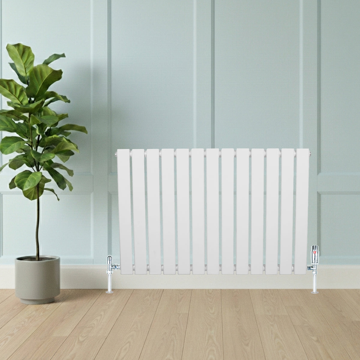 Flachheizkörper in Weiß, 600mm x 1190mm, mit vertikalen Lamellen und glänzenden Ventilen, vor einer hellen Wand und einem grünen Pflanzenarrangement