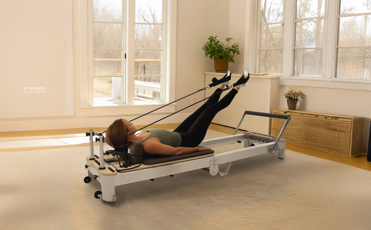 Eine Frau in Leggings und einem Tanktop trainiert auf einem Pilates-Reformer mit Widerstandsbändern in einem hellen Raum mit großen Fenstern