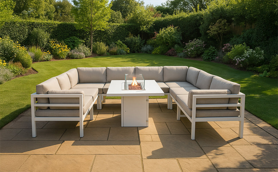 Moderne Gartenlounge mit beigem Polster, weißem Aluminiumrahmen und quadratischem Tisch mit Feuerstelle, umgeben von grüner Landschaft und bunten Blumen