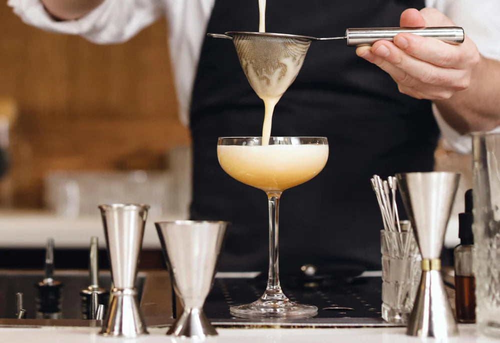 Ein Barkeeper gießt einen cremigen Cocktail durch ein feines Sieb in ein elegantes Glas, umgeben von Messbechern und Barutensilien