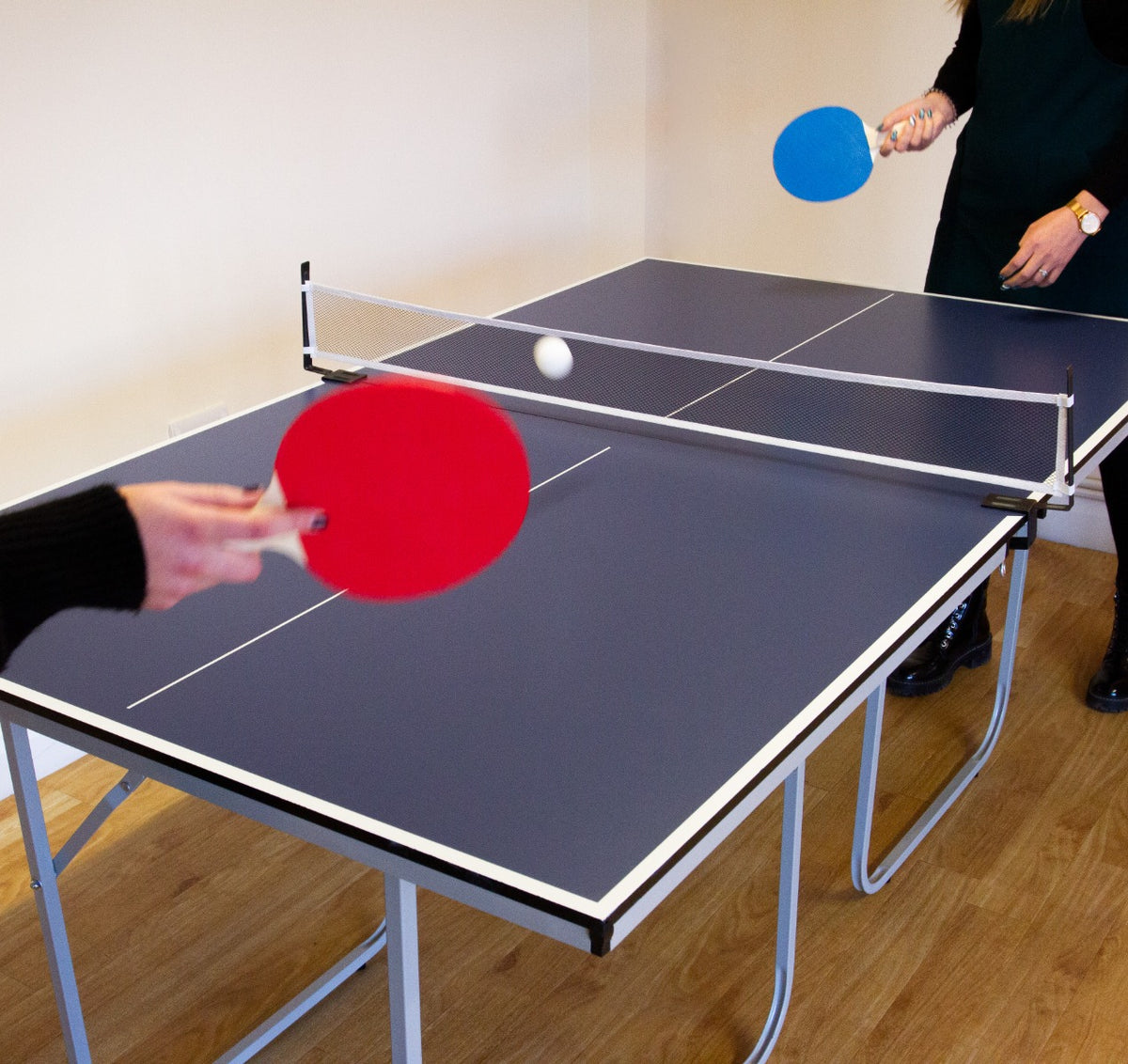 Klappbare Tischtennisplatte in Blau mit weißen Linien, zwei Spieler halten rote und blaue Schläger, Ball fliegt über das Netz