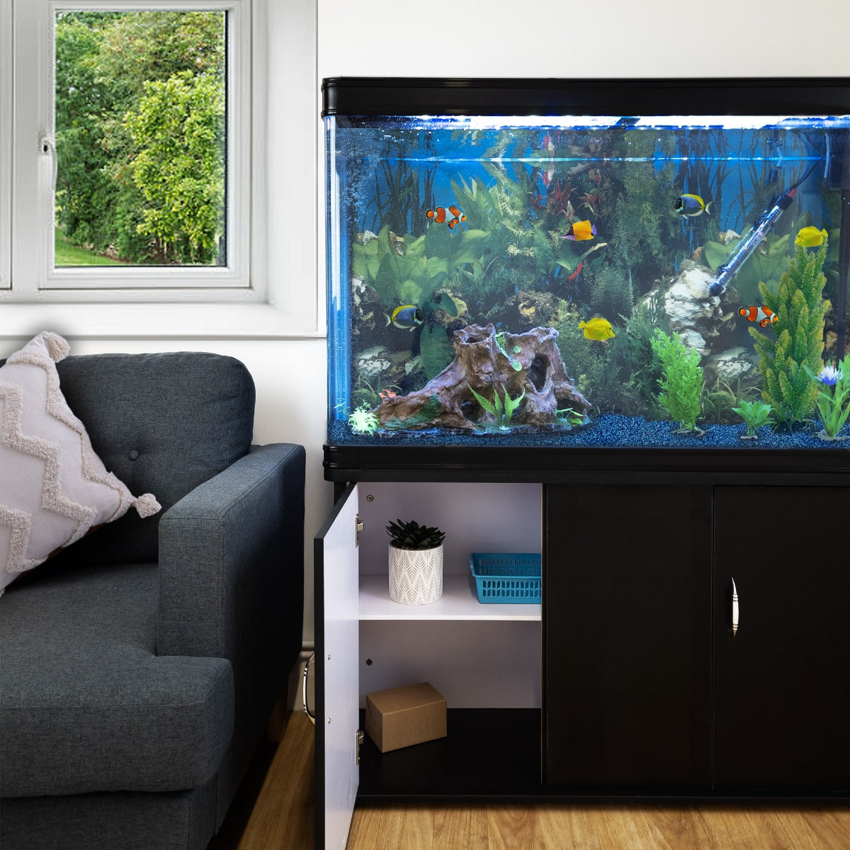 Schwarzes 300L Aquarium-Set mit bunten Fischen, lebendiger Pflanzenwelt und Kiesboden, platziert neben einem gemütlichen grauen Sofa und Fenster mit Ausblick