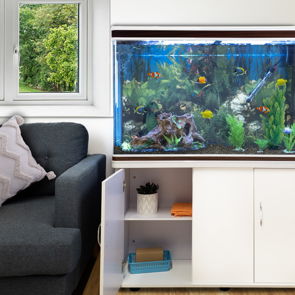 Aquarium Komplettset mit weißem Unterschrank, schwarzem Kies und bunten Fischen, umgeben von grünen Pflanzen und einem gemütlichen Wohnzimmer mit Sofa