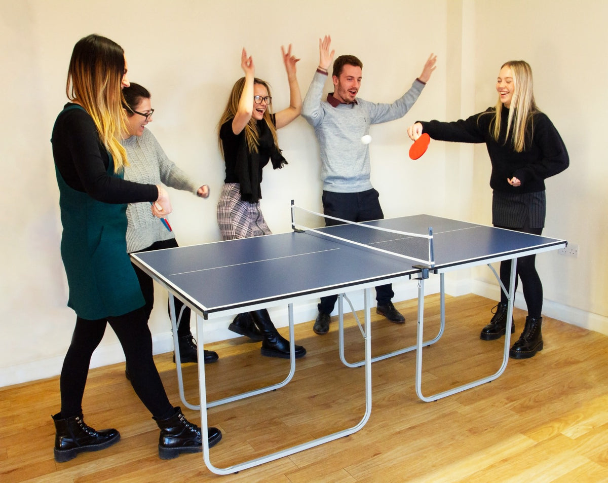 Fünf Personen spielen begeistert Tischtennis an einer klappbaren blauen Tischtennisplatte aus Metall mit einem hellen Holzfußboden