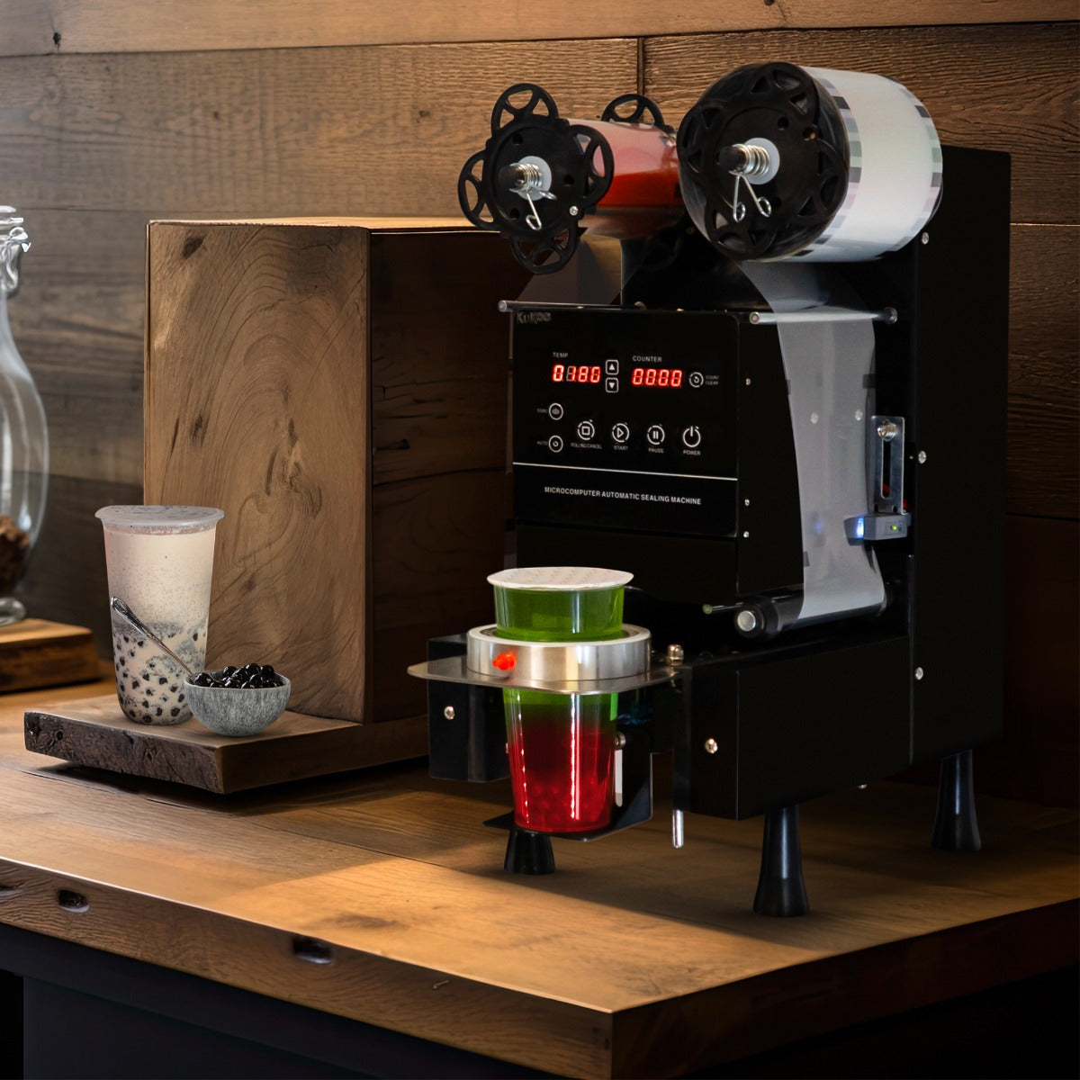 Vollautomatische Bechersiegelmaschine in Schwarz mit digitalen Anzeigen, daneben ein Becher mit bunten Schichten und Zutaten auf einem Holzregal