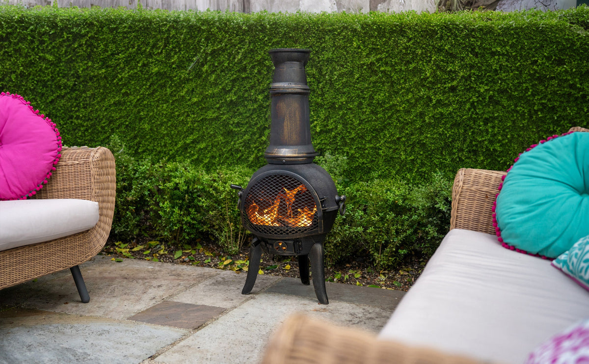 Gusseisen-Terrassenofen mit flammendem Feuer, umgeben von grünen Hecken und gemütlichen Rattanmöbeln mit bunten Kissen in Pink und Türkis