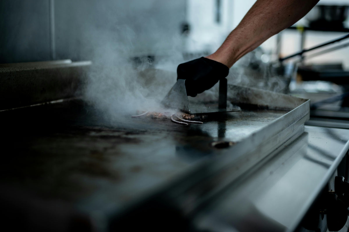Ein Koch mit schwarzer Handschuh greift mit einem Spachtel nach gegrilltem Fleisch auf einer glänzenden, heißen Elektro-Griddleplatte, umgeben von Dampf