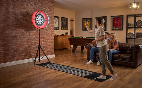 Eine Frau wirft einen Dartpfeil auf ein freistehendes Dartboard, während zwei Personen auf einer Couch im Hintergrund zuschauen, umgeben von Holz- und Ziegelwänden
