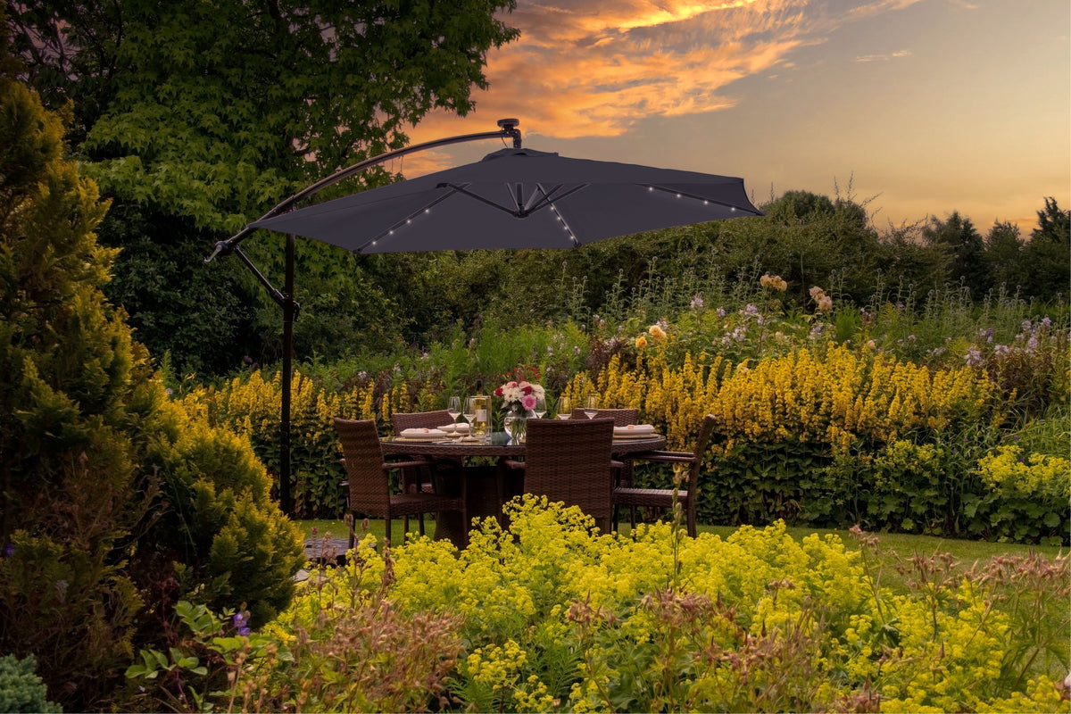Schwarzer Sonnenschirm mit LED-Lichtern über einem stilvollen Gartentisch, umgeben von bunten Blumen und üppigem Grün im Abendlicht