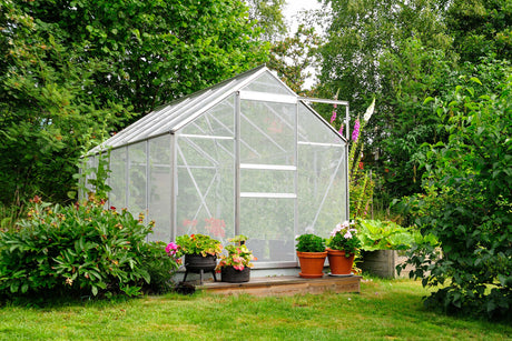 Ein modernes Gewächshaus aus klarem Glas, umgeben von bunten Blumen und Pflanzen in Töpfen, auf einer grünen Wiese im Frühling