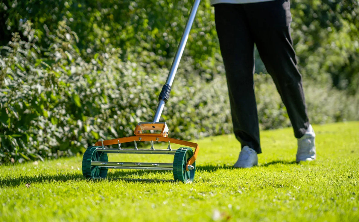 Ein Rasenmäher mit orangefarbenem Gestell und grünen Rädern wird über saftiges, grünes Gras geschoben, während eine Person in dunkler Kleidung daneben steht