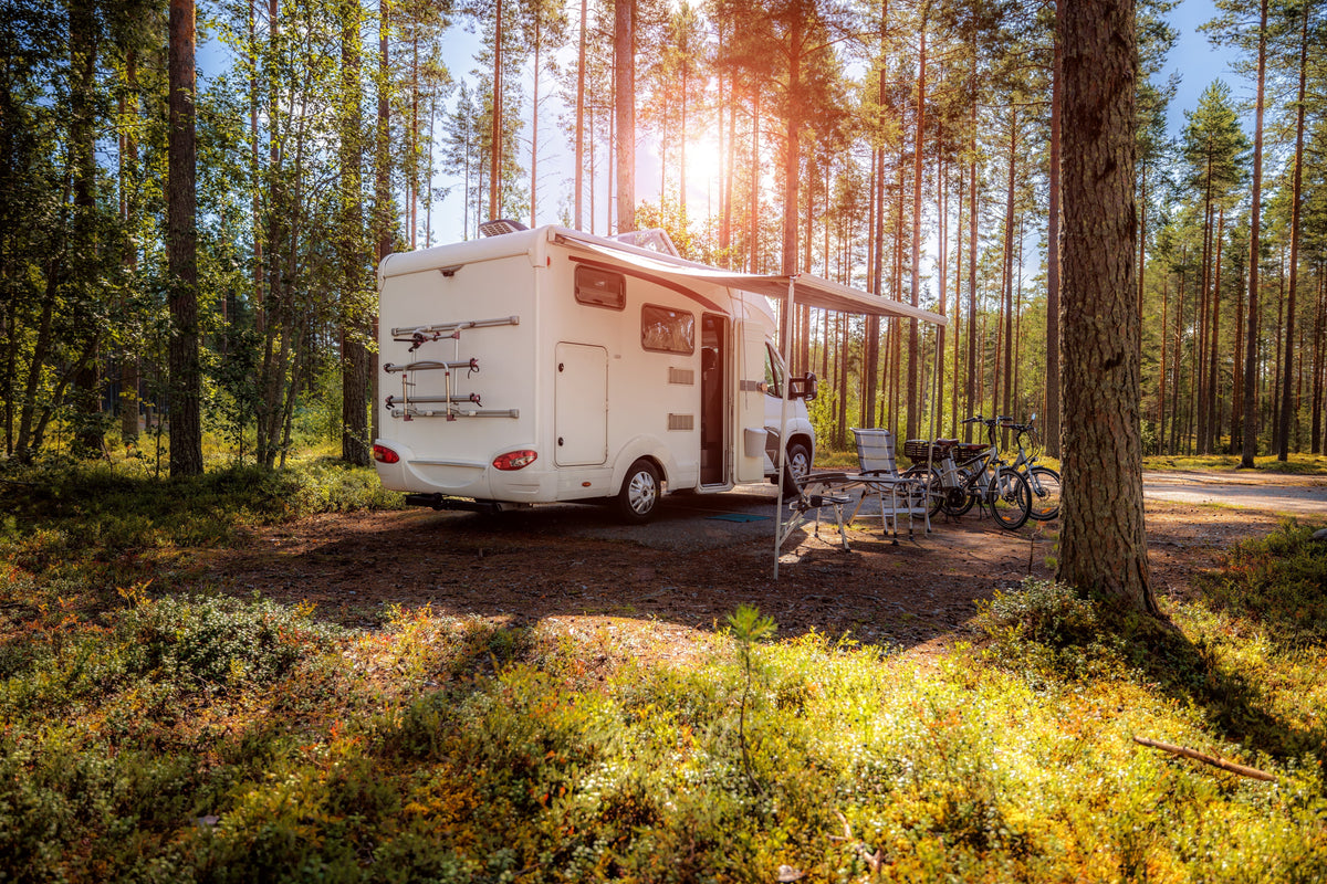 Ein weißer Wohnwagen steht in einem sonnigen Wald, umgeben von grünen Bäumen und buntem Moos, mit einem Fahrrad und Gartenmöbeln daneben